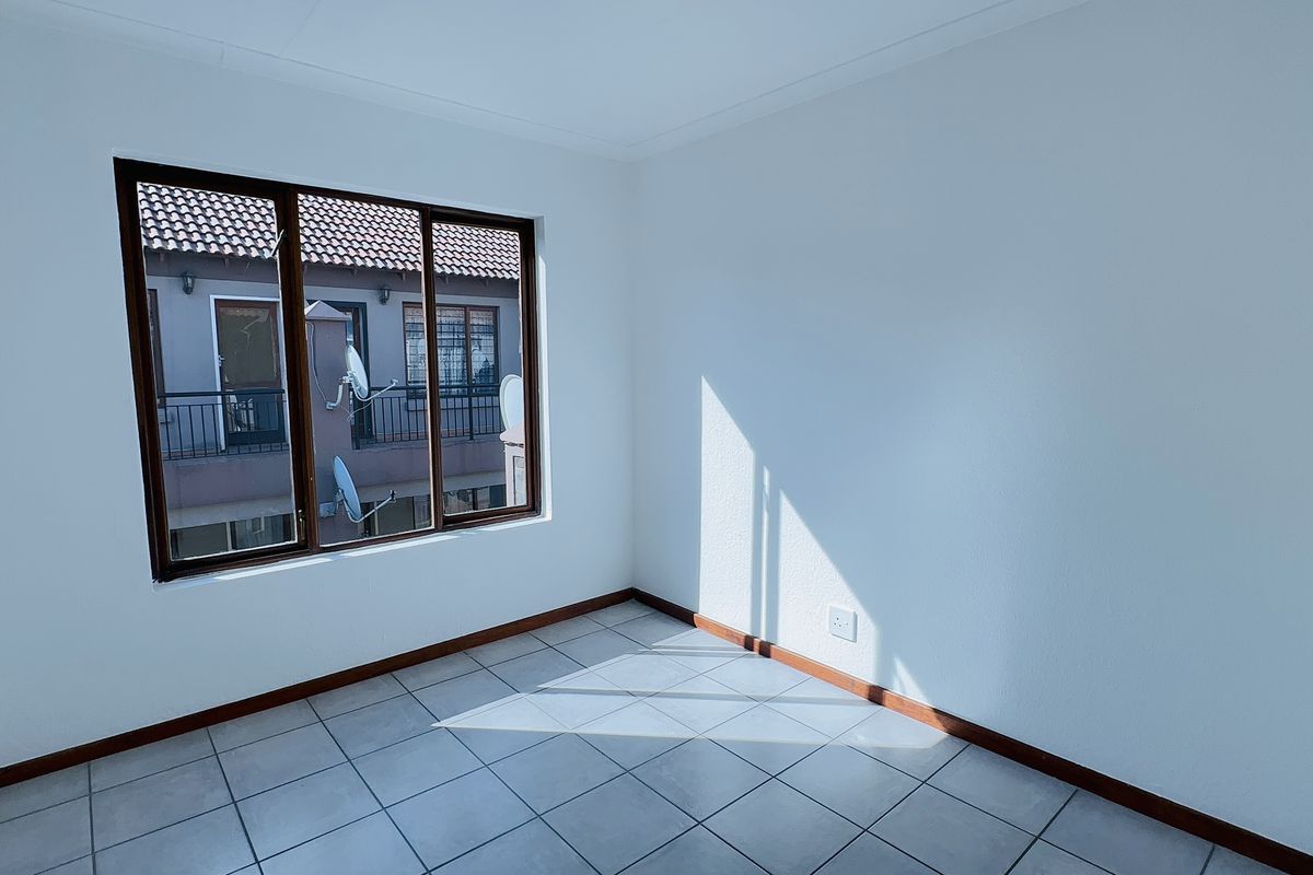 A bright, empty bedroom featuring neutral tiled flooring, clean white walls, and timber-framed windows overlooking the complex exterior.