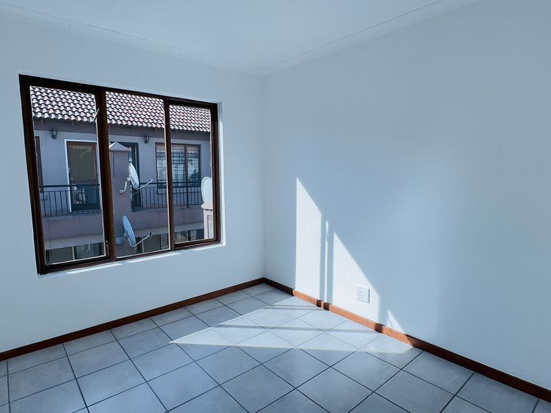 A bright, empty bedroom featuring neutral tiled flooring, clean white walls, and timber-framed windows overlooking the complex exterior.