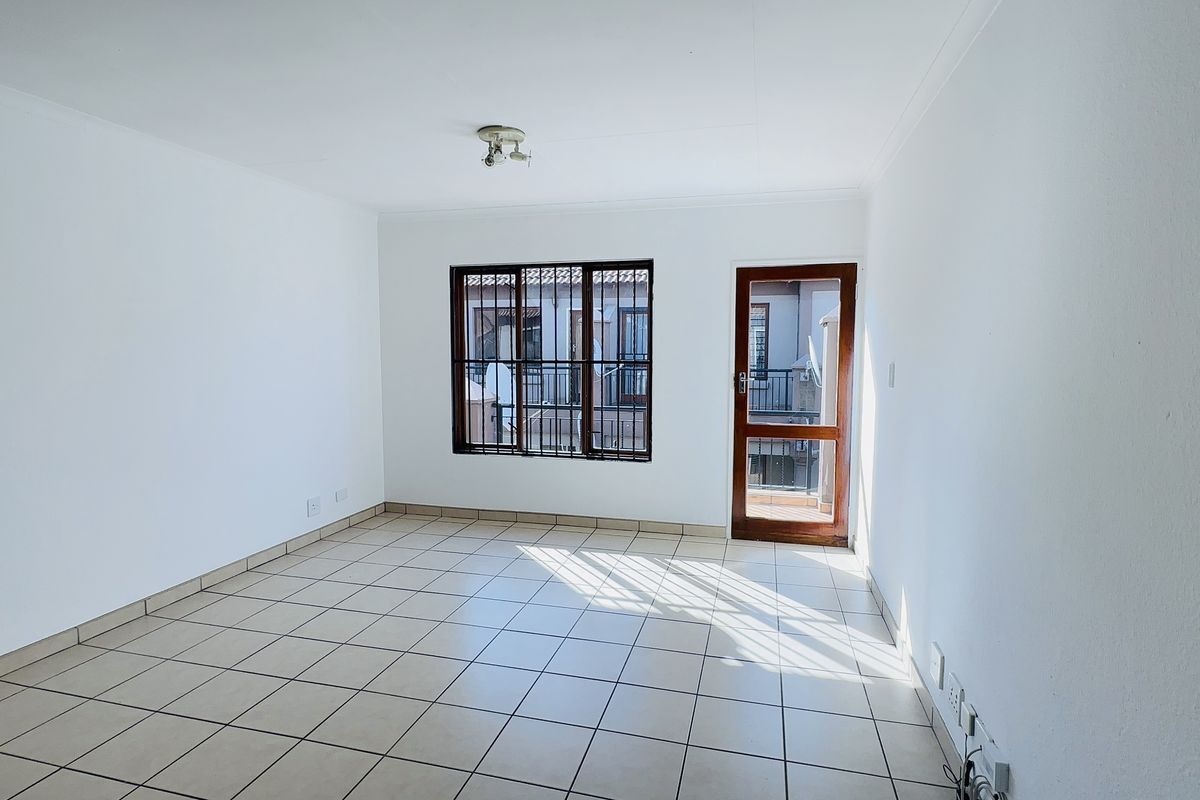 A bright, empty living room featuring neutral tiled flooring, a security-barred window, and a timber-framed door leading to an outdoor area.