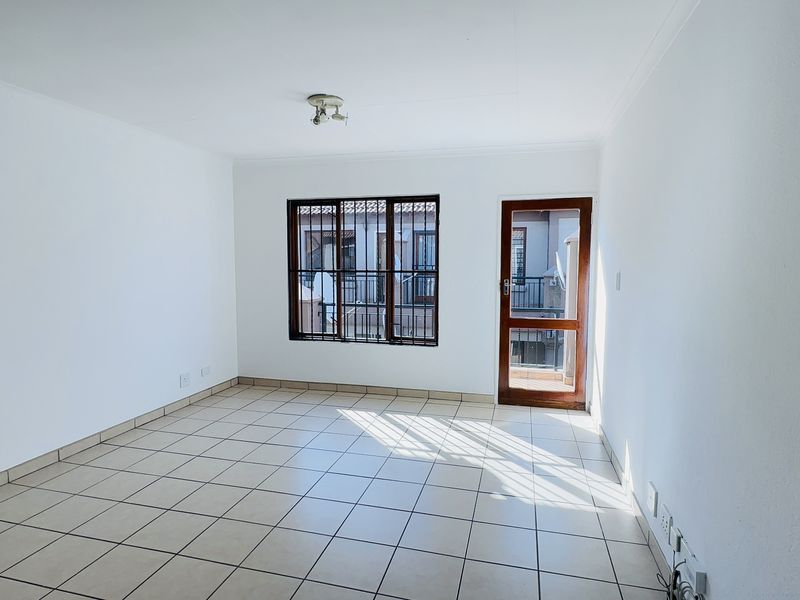 A bright, empty living room featuring neutral tiled flooring, a security-barred window, and a timber-framed door leading to an outdoor area.