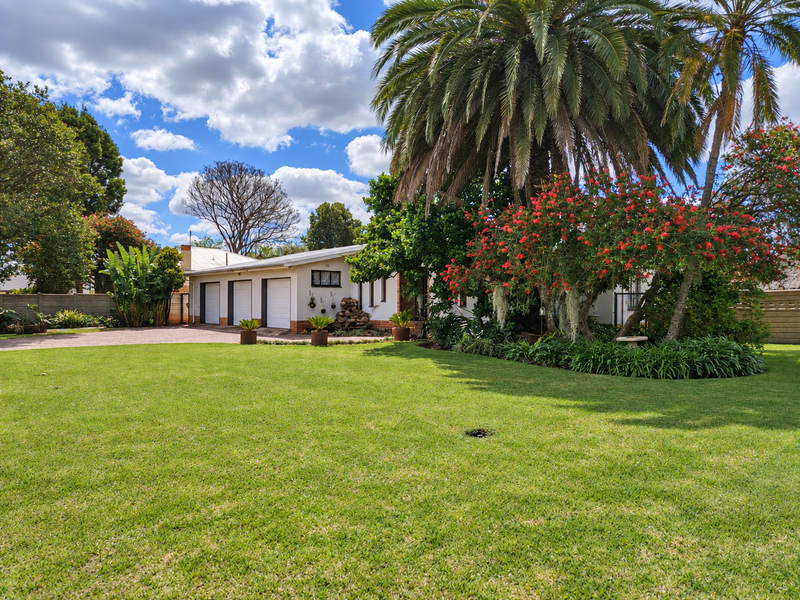 A well-maintained front exterior view featuring a spacious lawn, mature landscaping, and a triple garage with a paved driveway.