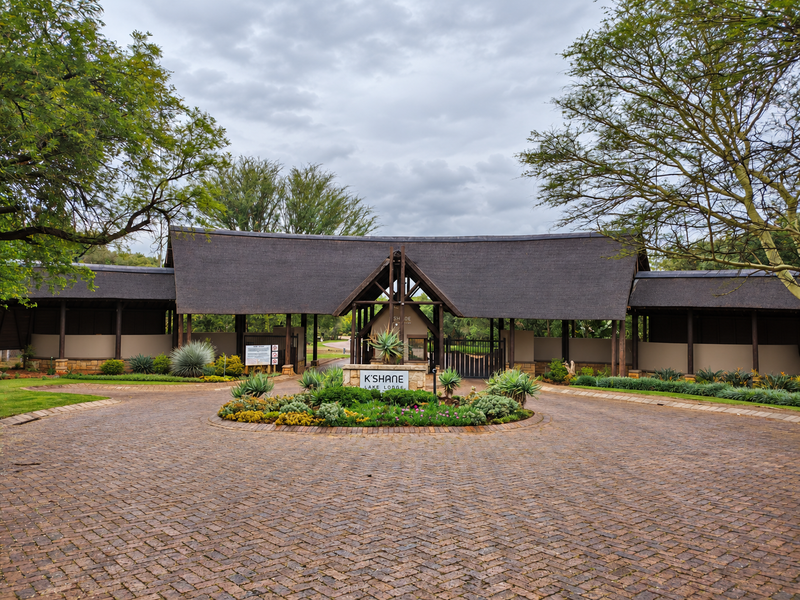 The grand entrance to K'Shane Lake Lodge featuring a distinctive thatched roof structure, brick paved driveway, and well-maintained landscaped gardens.