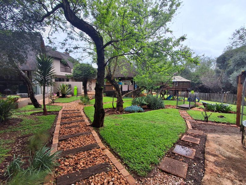 A spacious rear garden featuring a landscaped lawn, paved pathways, mature trees, and a thatched roof dwelling with a wooden play structure.