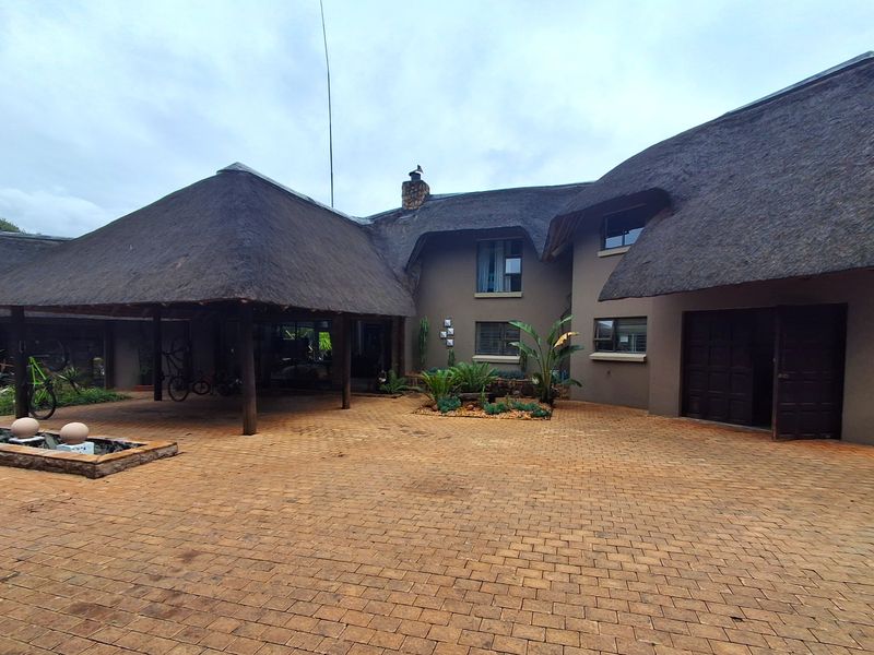 Spacious front exterior featuring a traditional thatch roof, extensive paved driveway, covered patio area, and a stone water feature.
