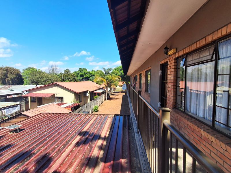 An elevated outdoor balcony walkway featuring brick wall finishes, metal railings, and views over the adjacent property and corrugated metal roofing.