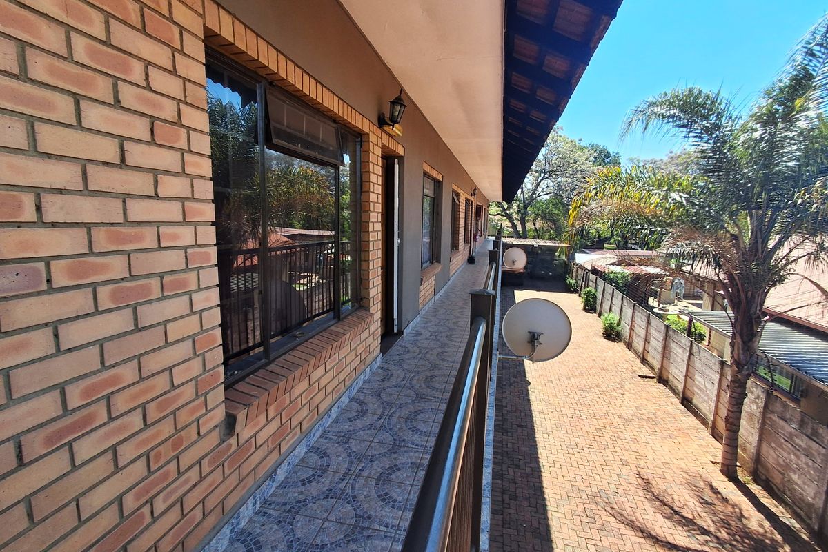 A long, covered balcony featuring brick exterior walls, tiled flooring, and a metal railing, overlooking the side yard and paved driveway area.