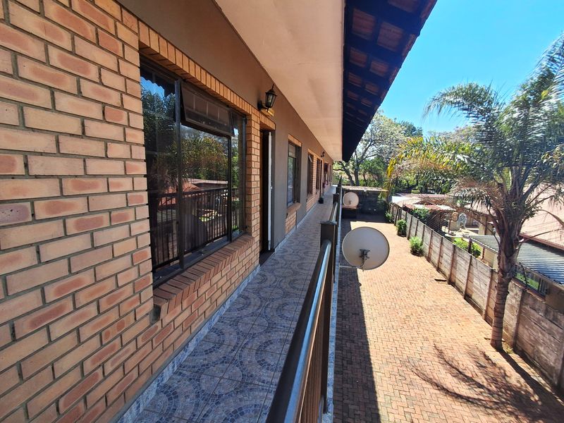 A long, covered balcony featuring brick exterior walls, tiled flooring, and a metal railing, overlooking the side yard and paved driveway area.