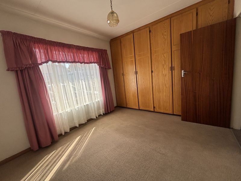 A spacious bedroom featuring extensive built-in wooden cupboards, neutral carpeting, and natural light through a large window.