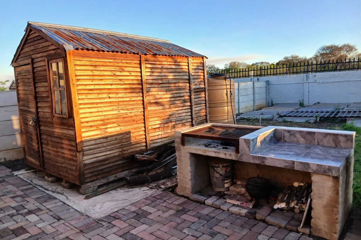 Rear garden area featuring a timber storage shed, a built-in brick braai with tiled prep surface, and a water storage tank on a paved patio.