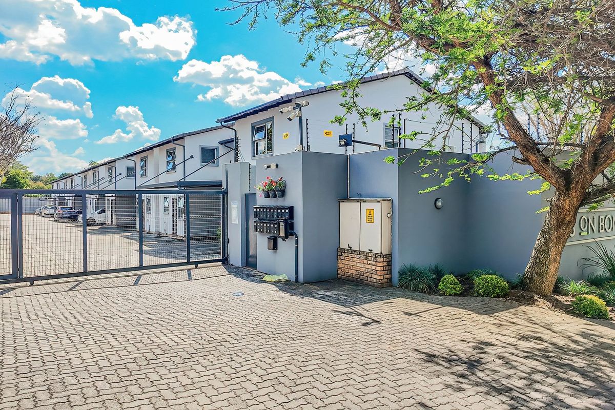 Front entrance view of the townhouse complex featuring a secure gated access point, paved driveway, and perimeter wall with electric fencing.