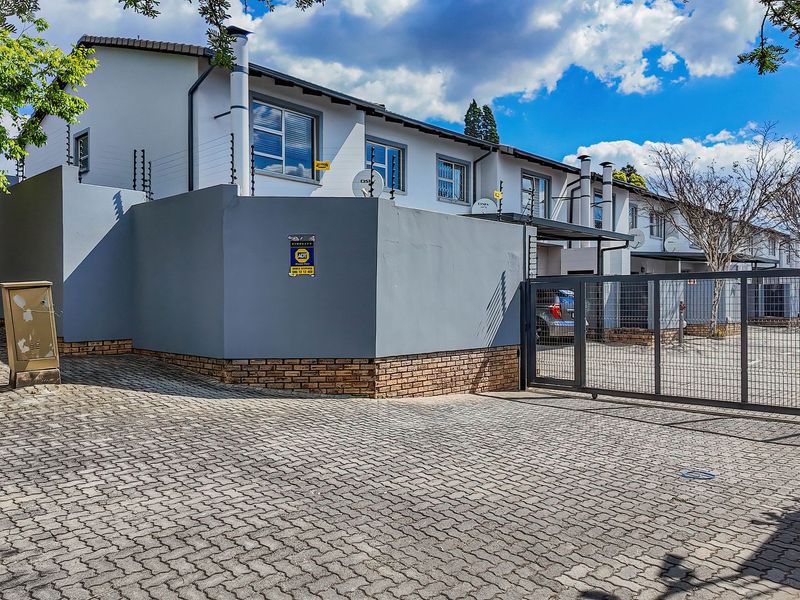 Front exterior view of a townhouse complex featuring a paved driveway, secure gated entrance, and perimeter wall with electric fencing.