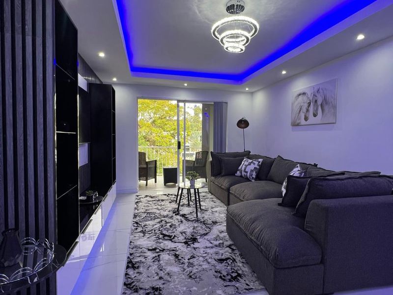 Modern living room featuring a recessed ceiling with ambient LED lighting, sleek tiled floors, and a contemporary slatted wall feature leading to a balcony.