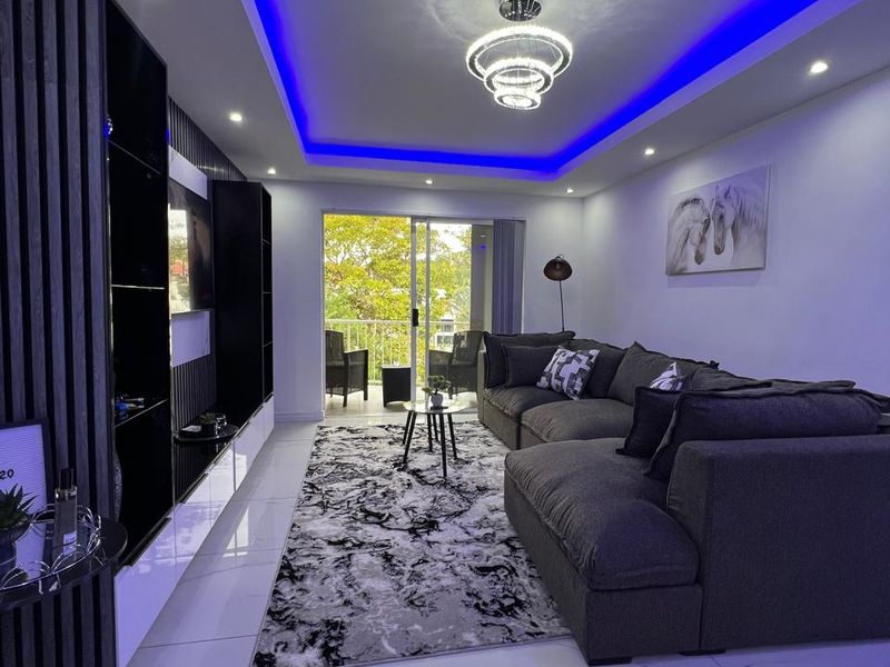 Modern living room featuring a built-in media wall, recessed ceiling lighting with blue LED accents, and large sliding glass doors leading to a balcony.