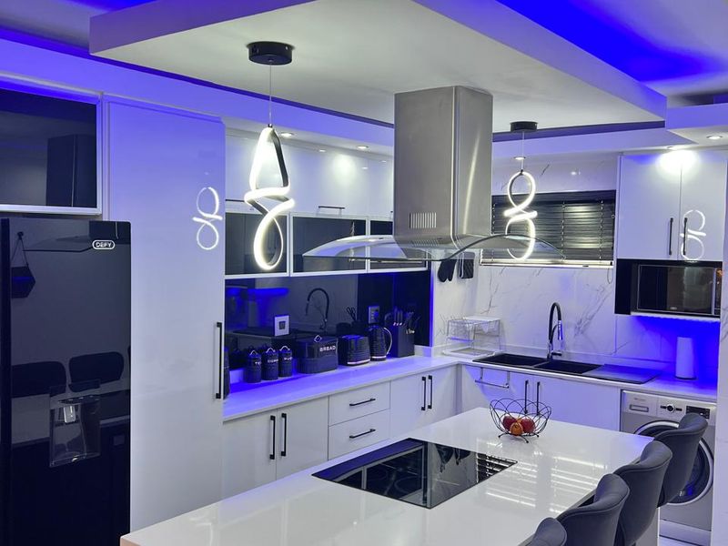 Modern kitchen featuring a central island with integrated hob, sleek white cabinetry, stone countertops, and contemporary LED accent lighting.