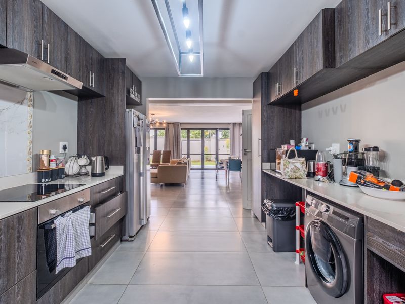 Modern galley-style kitchen featuring dark wood cabinetry, stone countertops, integrated appliances, and tiled flooring, leading into an open-plan living area.