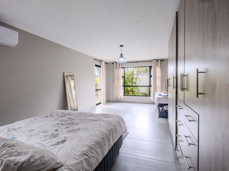 Spacious bedroom featuring modern built-in cupboards, tiled flooring, and an air conditioning unit, with large windows providing natural light and garden views.