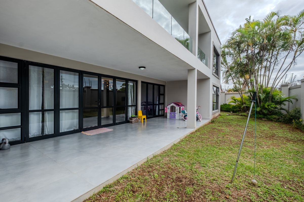 A spacious covered patio featuring large aluminum-framed glass doors, tiled flooring, and a modern glass balustrade, overlooking a private garden area.