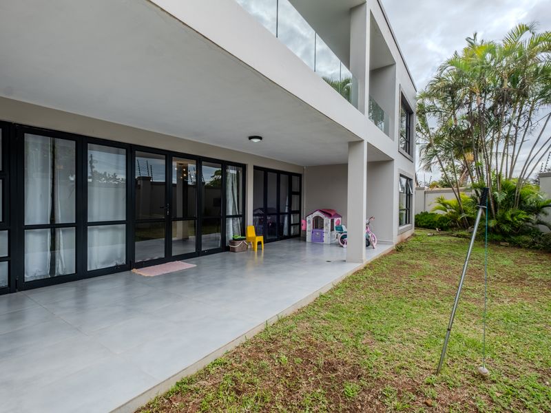 A spacious covered patio featuring large aluminum-framed glass doors, tiled flooring, and a modern glass balustrade, overlooking a private garden area.