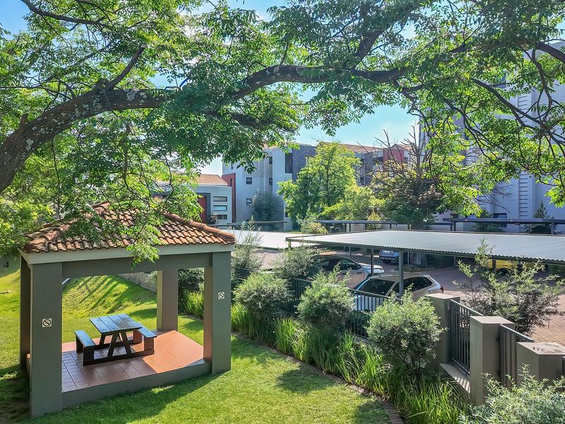 A well-maintained communal garden area featuring a covered gazebo, paved parking carports, and established landscaping within the apartment complex grounds.