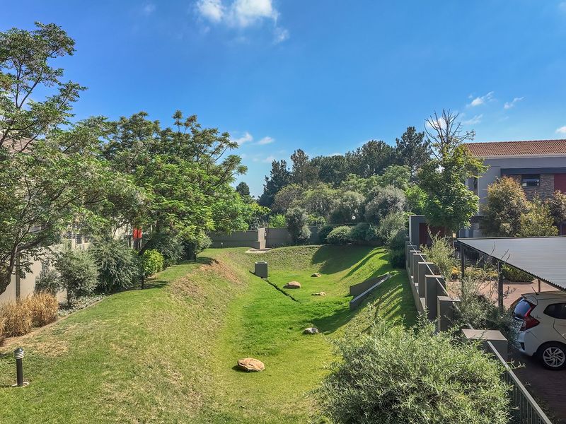 A well-maintained communal garden area featuring landscaped lawns, mature trees, and a covered carport structure within a residential apartment complex.