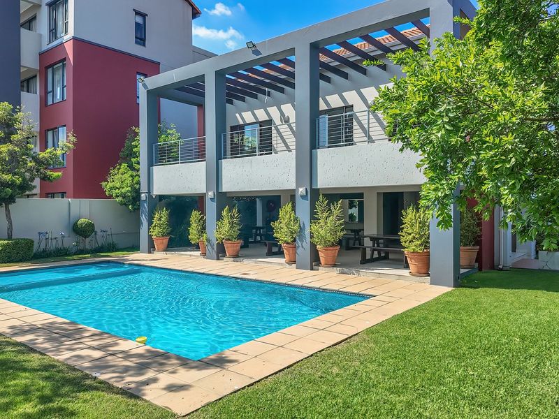 A well-maintained outdoor area featuring a swimming pool, paved deck, and a covered patio with a pergola, set within a landscaped garden.