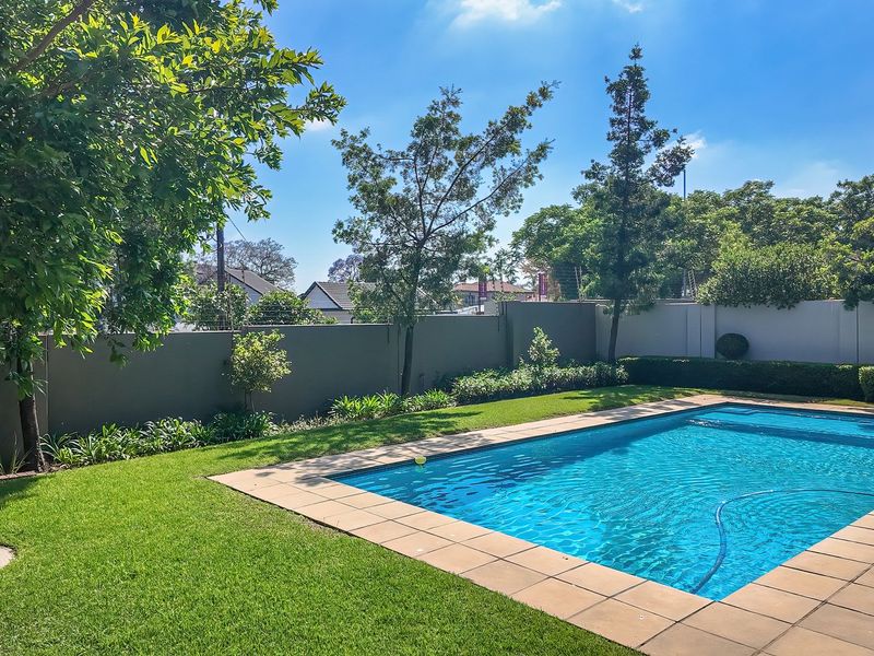 A well-maintained outdoor pool area featuring a clear swimming pool, tiled surround, and a landscaped garden enclosed by a solid perimeter wall.