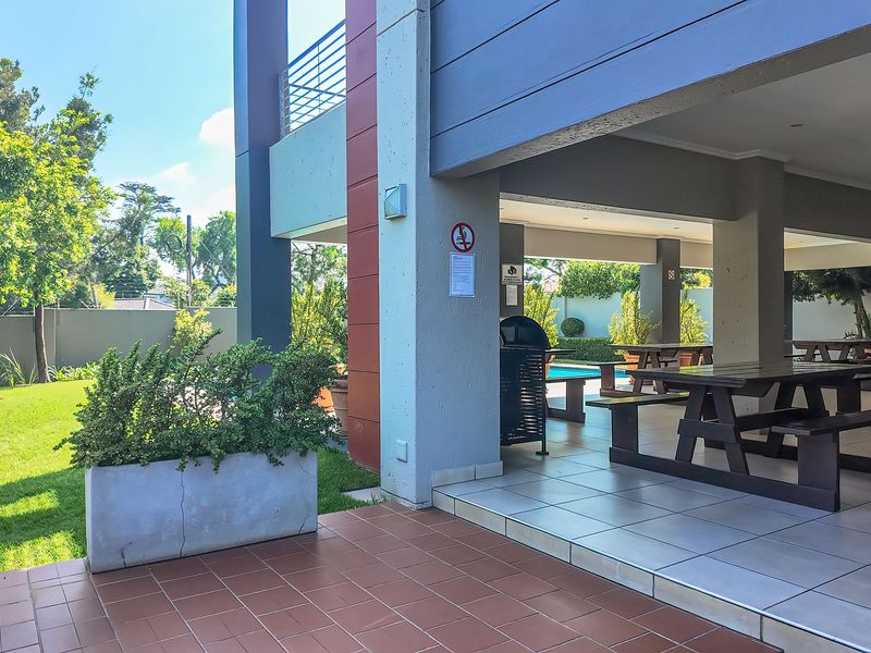 A spacious covered outdoor patio area featuring tiled flooring, concrete structural columns, and integrated planter boxes, overlooking a landscaped garden and pool area.