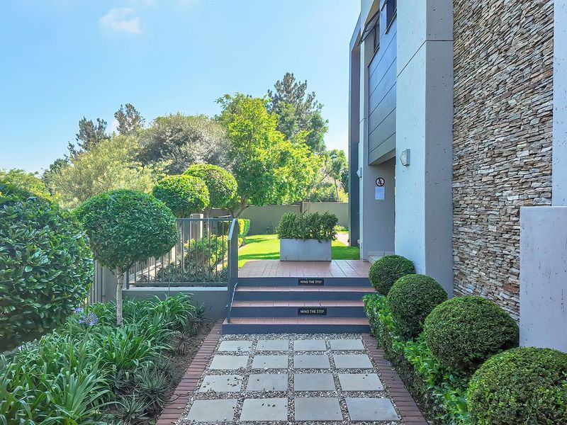 A well-maintained front entrance featuring a paved walkway, stone-clad exterior wall, and manicured garden landscaping leading to the building entrance.