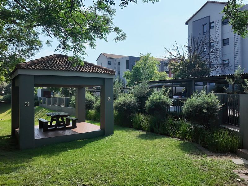 A well-maintained communal garden area featuring a covered gazebo with tiled flooring, surrounded by landscaped lawns and established shrubbery within the apartment complex.