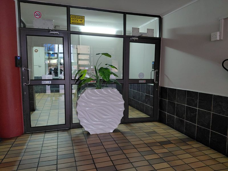 A functional apartment building entrance lobby featuring tiled flooring, dark tiled wall accents, and secure glass-panelled aluminium doors with integrated access control.
