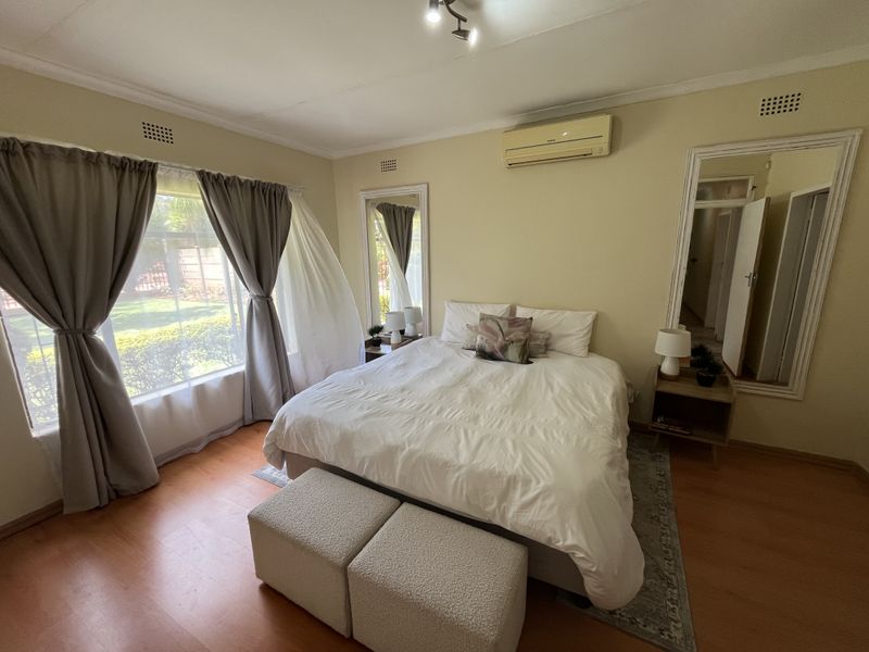 A well-lit bedroom featuring laminate flooring, an air conditioning unit, and large windows overlooking the garden.