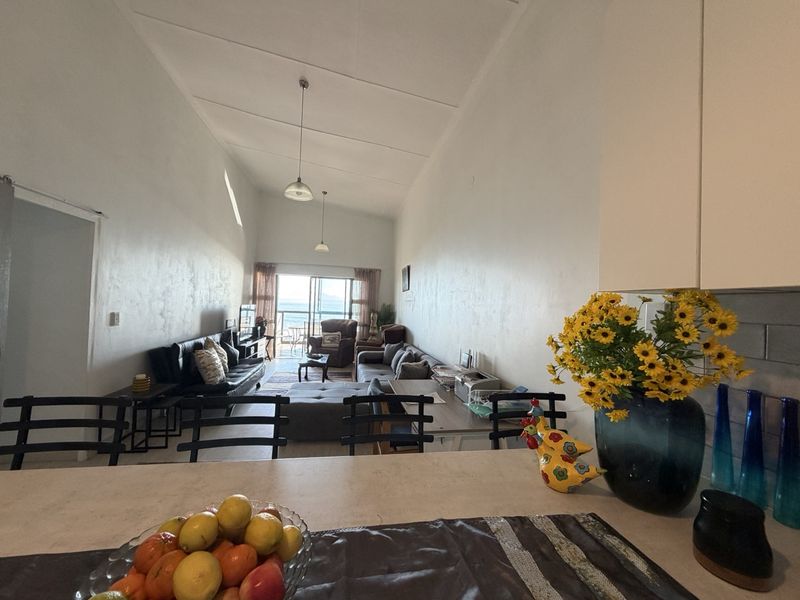 Spacious open-plan living and kitchen area featuring a vaulted ceiling, kitchen island, and sliding glass doors leading to a balcony with ocean views.