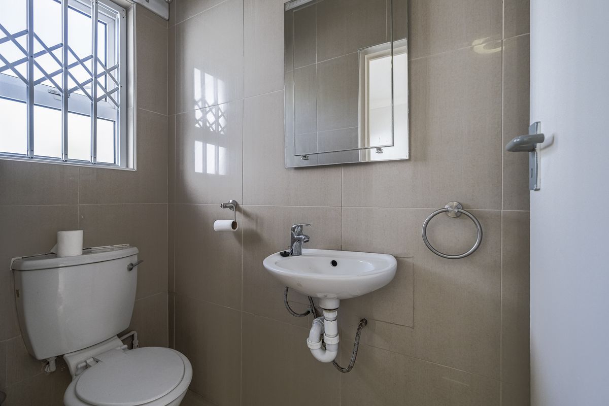 A clean and functional guest bathroom featuring neutral wall tiles, a wall-mounted basin, a toilet, and a security-barred window.