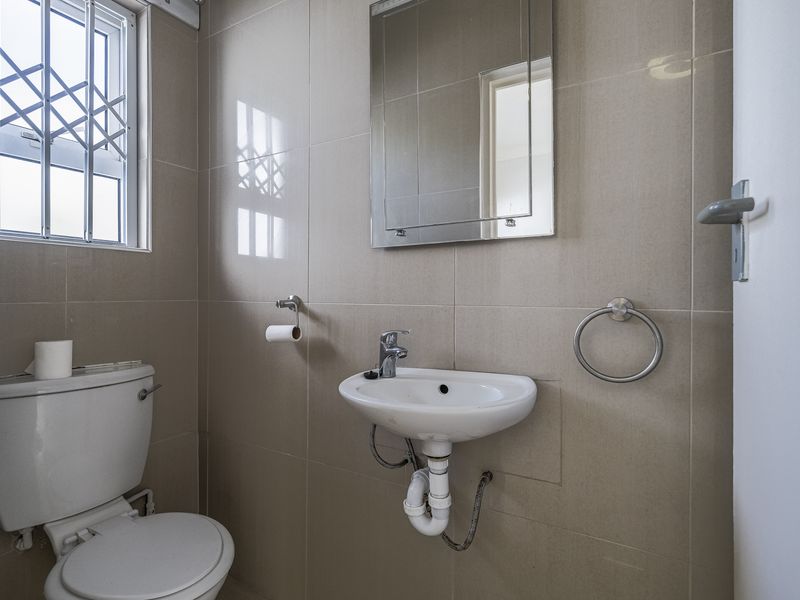 A clean and functional guest bathroom featuring neutral wall tiles, a wall-mounted basin, a toilet, and a security-barred window.