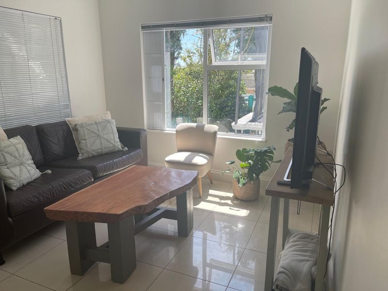A bright living room featuring tiled flooring and large windows with venetian blinds, offering a view of the garden.
