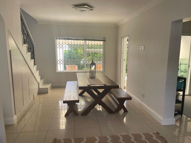 A bright dining area featuring tiled flooring, a staircase with a decorative wrought iron balustrade, and a window fitted with a security gate.