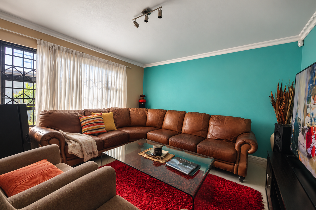 A comfortable living room featuring a vibrant teal accent wall, ceiling coving, and track lighting, with large windows fitted with security bars.