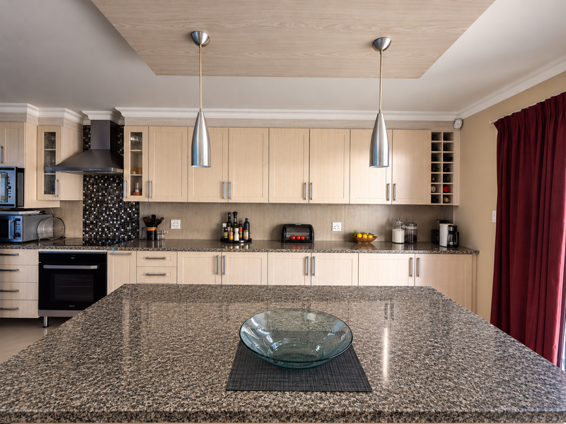 Modern kitchen featuring light wood cabinetry, granite countertops, a central island, mosaic backsplash, and integrated stainless steel appliances under a decorative drop ceiling.