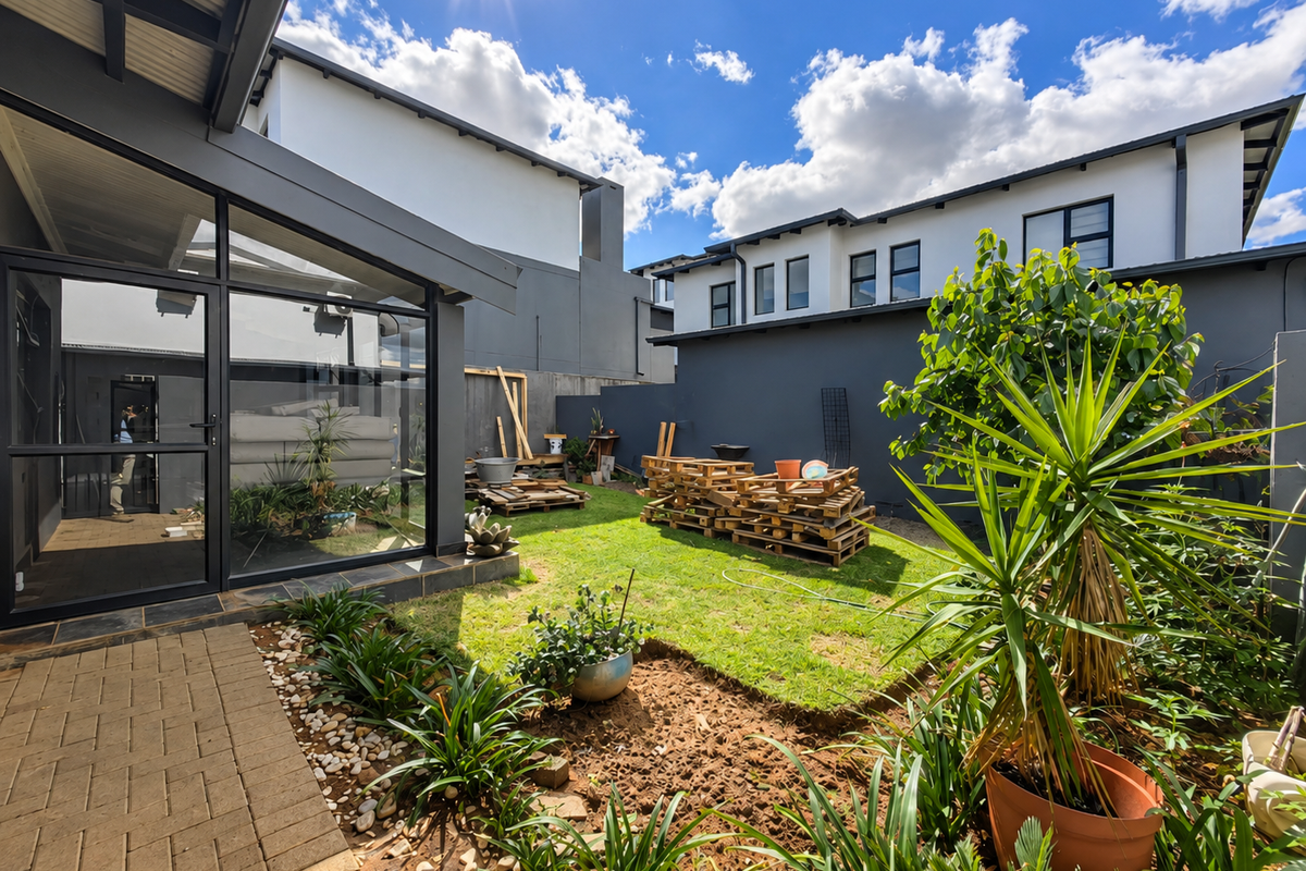 A rear garden area featuring a paved patio, lawn, and modern townhouse architecture with large glass doors and dark grey exterior finishes.