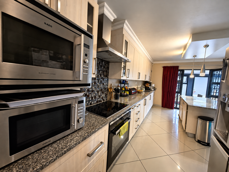 Modern kitchen featuring light wood cabinetry, granite countertops, mosaic backsplash, built-in appliances, and a central island, flowing into an open-plan living area.