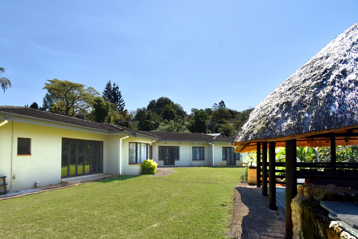 Rear view of the house featuring a spacious lawn, paved patio area, and a traditional thatched lapa structure.