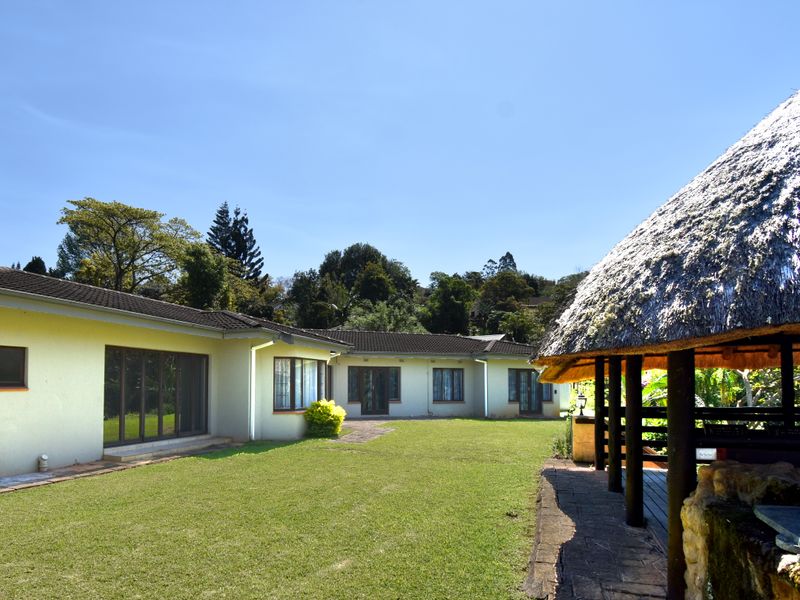 Rear view of the house featuring a spacious lawn, paved patio area, and a traditional thatched lapa structure.