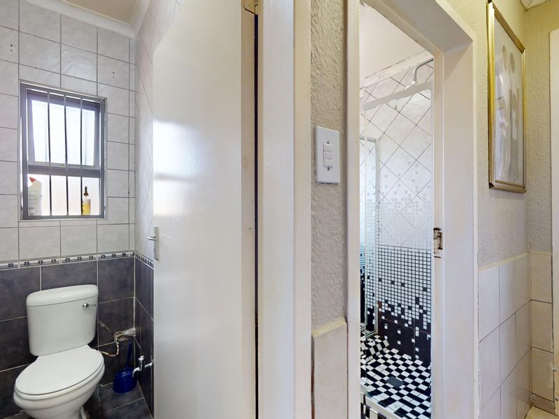 A functional bathroom featuring a toilet and a separate shower enclosure with contrasting tiled finishes and a window with security bars.