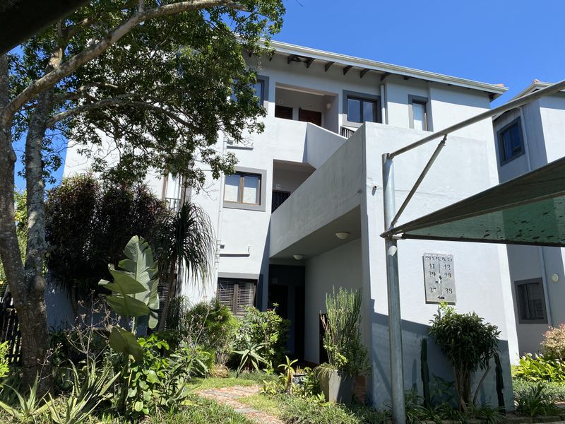 Exterior view of a multi-story apartment building featuring a covered walkway, paved entrance path, and established garden landscaping.