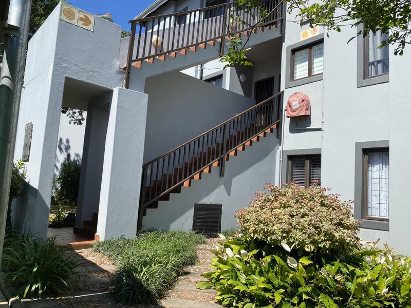 Exterior view of an apartment building featuring a paved walkway, landscaped garden, and an external staircase leading to the upper level entrance.