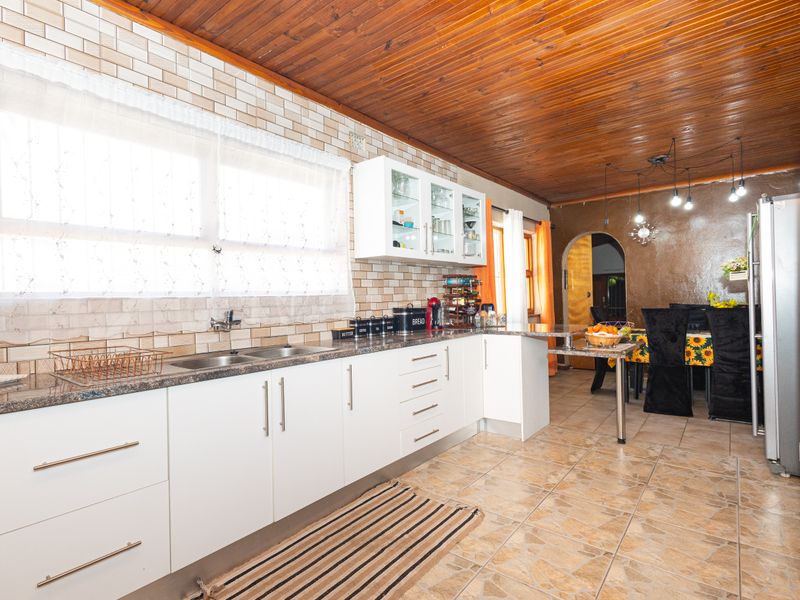 Open-plan kitchen and dining area featuring modern white cabinetry, stone-look countertops, tiled flooring, and a distinctive timber-clad ceiling.