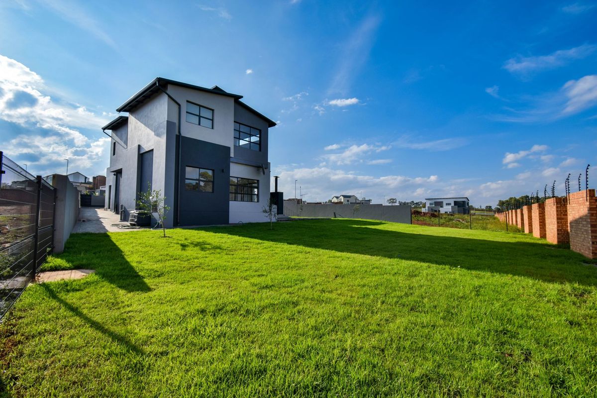 A view of the rear exterior of a modern two-story house featuring a spacious lawn, paved walkway, and perimeter fencing.