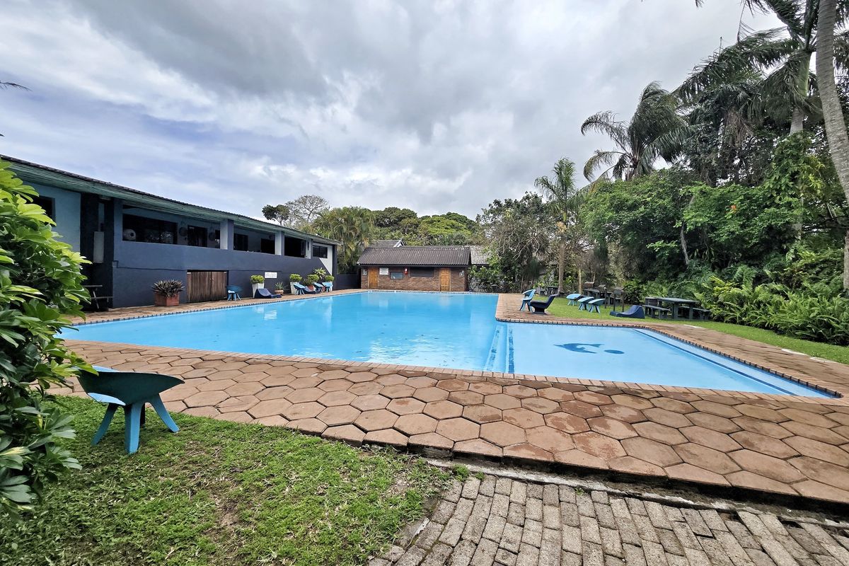 A large outdoor swimming pool area featuring paved surrounds, established garden landscaping, and adjacent brick outbuildings.