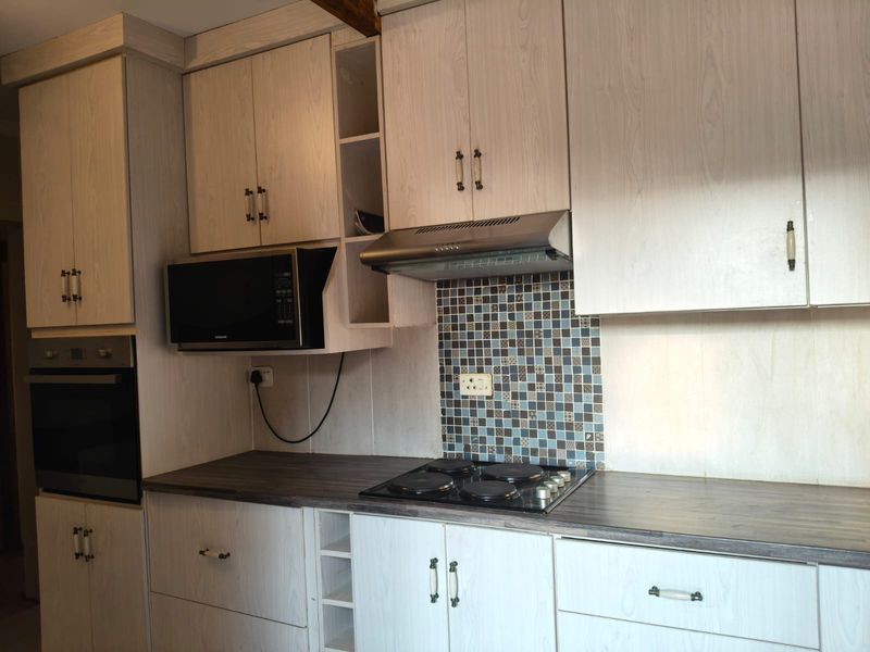 A functional kitchen featuring light wood-grain cabinetry, a dark laminate countertop, mosaic tile backsplash, and integrated appliances including an oven and electric hob.