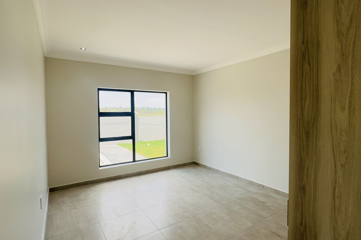 A bright, empty bedroom featuring neutral walls, tiled flooring, and a large aluminium-framed window providing natural light.